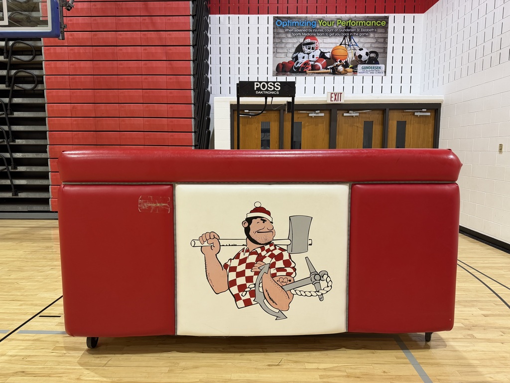 front side of Riverman scorers table that was used in the gym for volleyball and basketball games for the scorers to sit on. The front is a heavy plastic material that is on wheels with bar like seating spot, made of wood and gray carpet floor front is red and white with a riverman logo in the middle, the