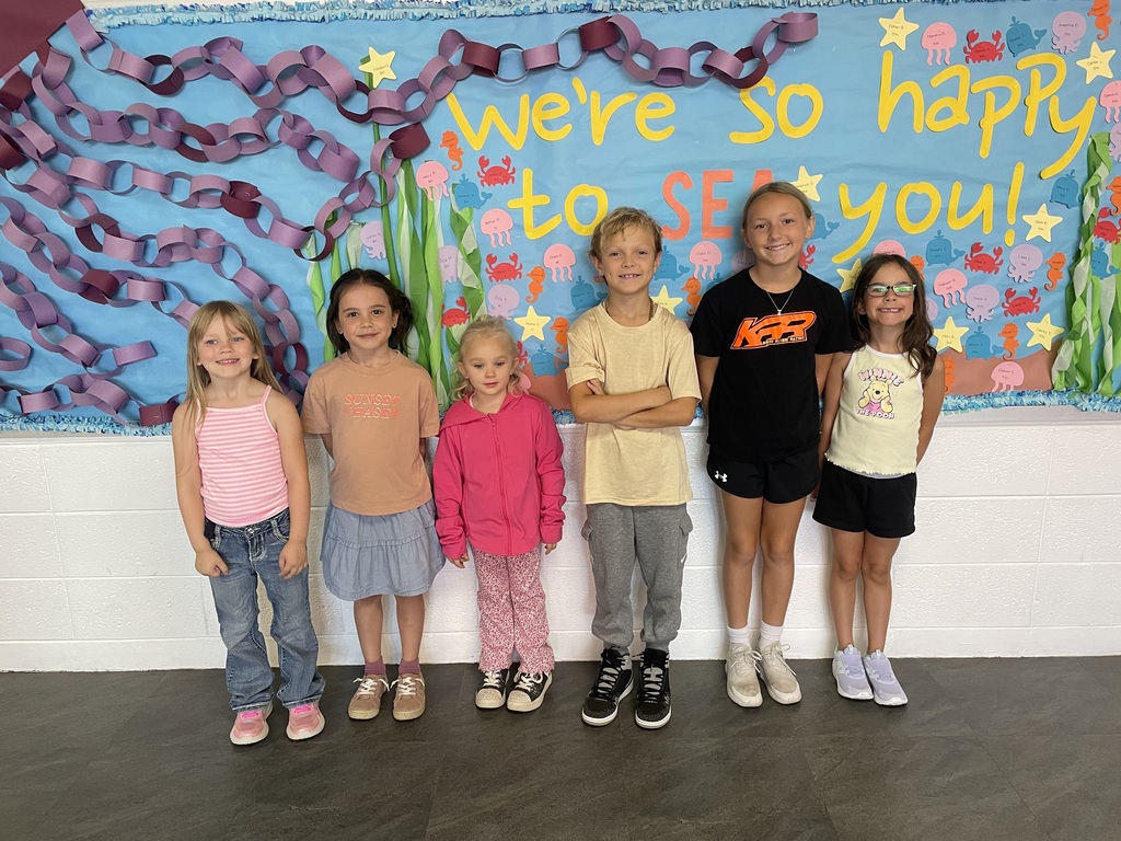 Six children stand side by side in front of a colorful ocean-themed bulletin board decorated with sea creatures, seaweed, stars, and paper chains. The display reads, “We’re so happy to see you!”