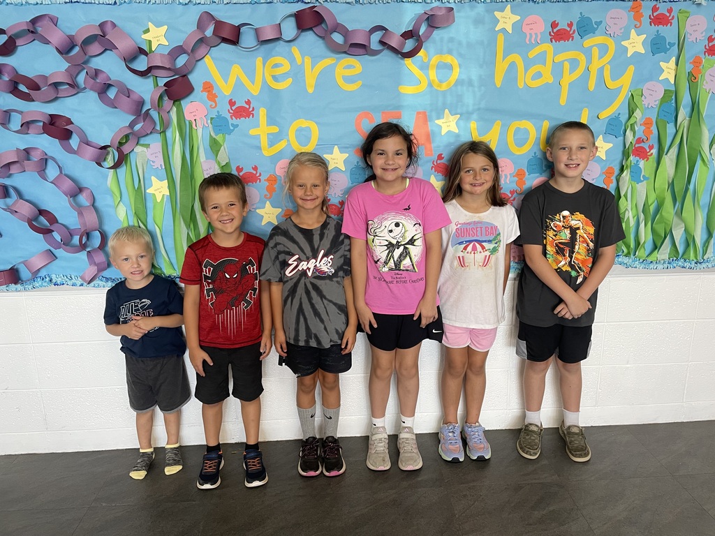 Six elementary-age children stand side by side in front of a colorful underwater-themed bulletin board decorated with sea creatures, seaweed, stars, and paper chains. The display reads, “We’re so happy to SEA you!”