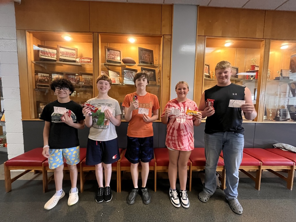 Five students stand together in front of a school trophy and memorabilia display case, holding small prizes and cards. They are smiling at the camera, with red auditorium-style chairs behind them.