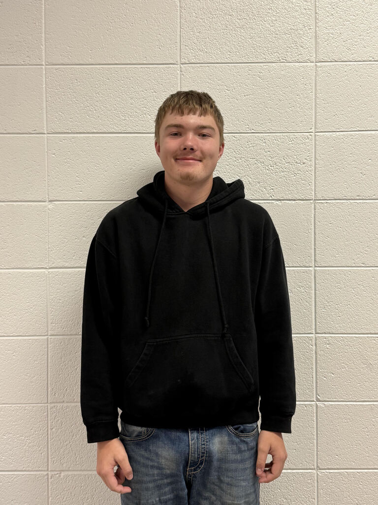 A student stands facing the camera in front of a white cinderblock wall, wearing a black hoodie and blue jeans.