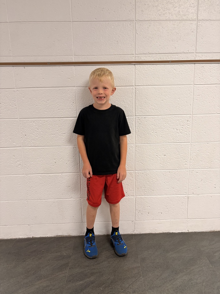 A young boy wearing a black shirt, red shorts, and blue shoes standing in front of a white wall.