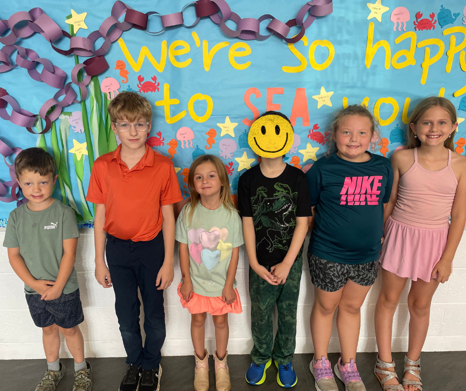 Six young students standing in a line side-by-side in front of a colorful, under-the-sea decorated classroom bulletin board that says "We're So Happy to SEA You!" in yellow and orange letters with paper chain seaweed. One student near the center has a digital yellow smiley face icon covering their face.