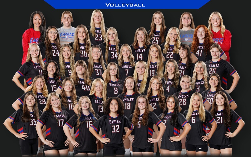 Group photo of the girls’ volleyball team posing together in black Eagles jerseys with red and blue accents. The players are arranged in several rows in front of a black background with a blue “VOLLEYBALL” banner at the top.