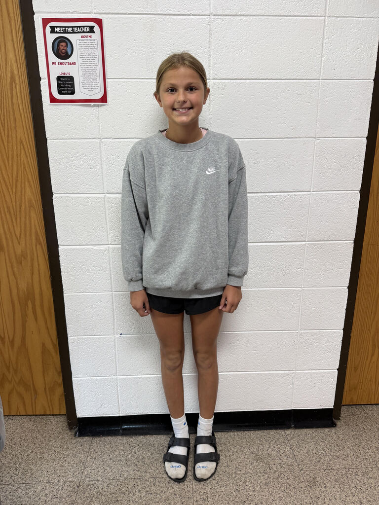 Student smiling and posing in a school hallway, wearing a gray Nike sweatshirt, black shorts, white socks, and black sandals. A “Meet the Teacher” poster for Mr. Engstrand is displayed on the wall behind them.