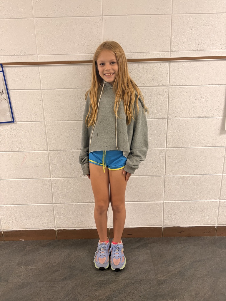 A young girl wearing a gray long-sleeved shirt, blue shorts, and purple sneakers standing in front of a white wall.