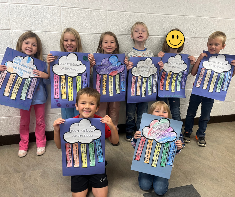 Eight young elementary school students stand in two rows against a painted cinder block wall, holding up personalized "be a rainbow of kindness" art projects. Five children stand in the back row, and two kneel or sit in the front. Each project consists of a large sheet of blue paper with a cloud cutout at the top labeled "be a rainbow of kindness," below which several colorful strips of paper hang down like a rainbow, each containing handwritten compliments and acts of kindness. One child's face in the back row is covered by a yellow smiley face emoji. The children are all smiling enthusiastically at the camera.