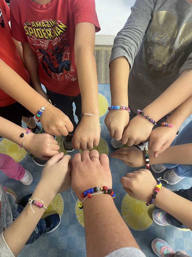 An overhead shot shows a group of young children and one adult gathering their arms together in a central circle to display colorful handmade pony bead bracelets. Each person wears a custom-made bracelet made of colorful plastic beads stringed on clear or white cord. The children wear a variety of casual clothing, including a red "Spider-Man" t-shirt, and stand on a blue carpet with yellow pattern accents.