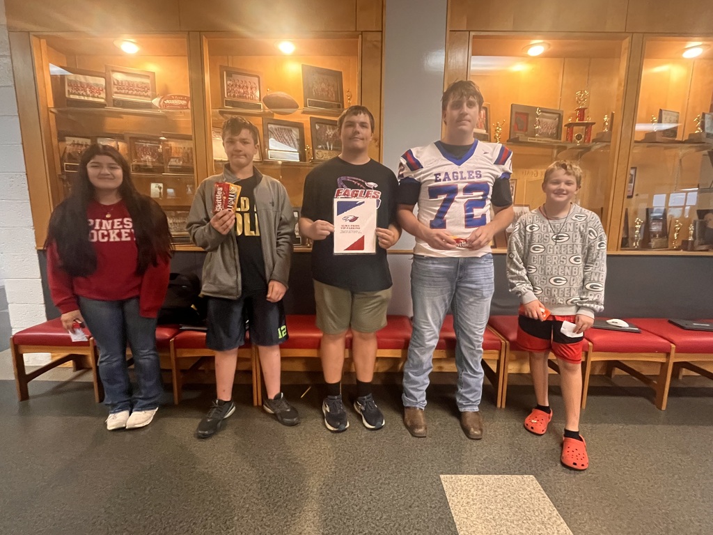 Five students stand together in front of a trophy display case. One student holds a candy, while another wears an Eagles football jersey. The group poses for a photo in a school hallway.