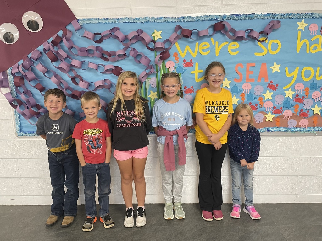 Six children stand side by side in front of a colorful ocean-themed school bulletin board decorated with paper chains, sea animals, stars, and the words “We’re so happy to SEA you.” The children are smiling and wearing casual clothing.