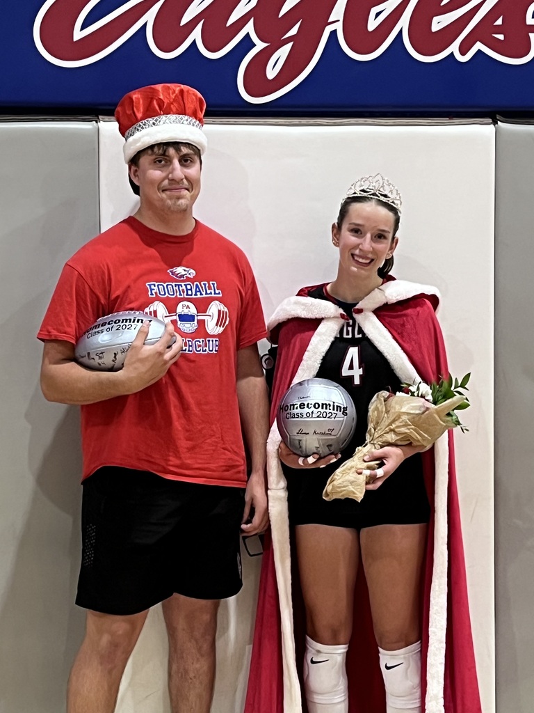 Max Crothers, the homecoming king, & Macy Stewart, the homecoming queen. Two students pose together at a school homecoming event. The student on the left wears a red football T-shirt, black shorts, and a red-and-white crown while holding a signed football. The student on the right wears a black volleyball uniform with the number 4, a red-and-white royal cape, and a tiara while holding a signed volleyball and a bouquet of flowers. A blue-and-red school banner is visible in the background.