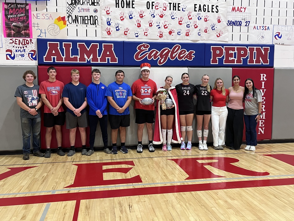 A group of 12 students poses on a school gymnasium floor beneath a large “ALMA Eagles PEPIN” sign. The students are dressed in a mix of school sports uniforms and casual clothing. Two students in the center wear homecoming crowns and hold signed footballs and volleyballs, with one also wearing a red-and-white cape and holding flowers. Red, blue, and white senior posters and handprint decorations cover the wall behind the group.