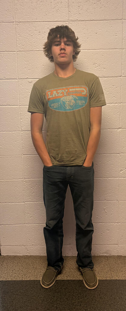 A student stands against a beige cinder-block wall, wearing an olive-green graphic T-shirt, dark jeans, and brown slip-on shoes. He has medium-length brown hair and stands with his hands in his pockets.