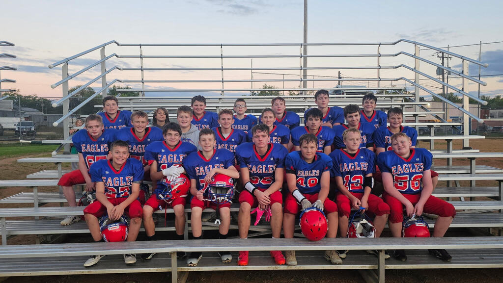 A youth football team of about 20 players poses together on outdoor bleachers. The players wear blue Eagles jerseys with red pants, and several hold or have red football helmets beside them. The team is grouped in three rows with a football field, fence, and evening sky in the background.