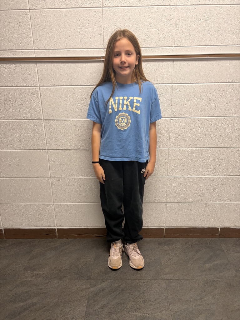 A young girl stands smiling in front of a light-colored cinderblock wall. She is wearing a blue Nike T-shirt, black athletic pants, and light-colored sneakers, with her hands at her sides. A wooden rail runs horizontally along the wall behind her.