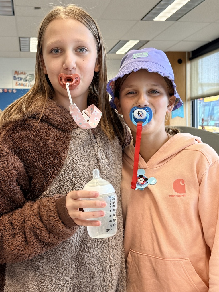   Two children pose together indoors at school. The child on the left wears a fuzzy brown-and-gray outfit, holds a baby bottle, and has a pink pacifier. The child on the right wears a peach hoodie and a purple sun hat, with a blue pacifier and a red pacifier clip. 