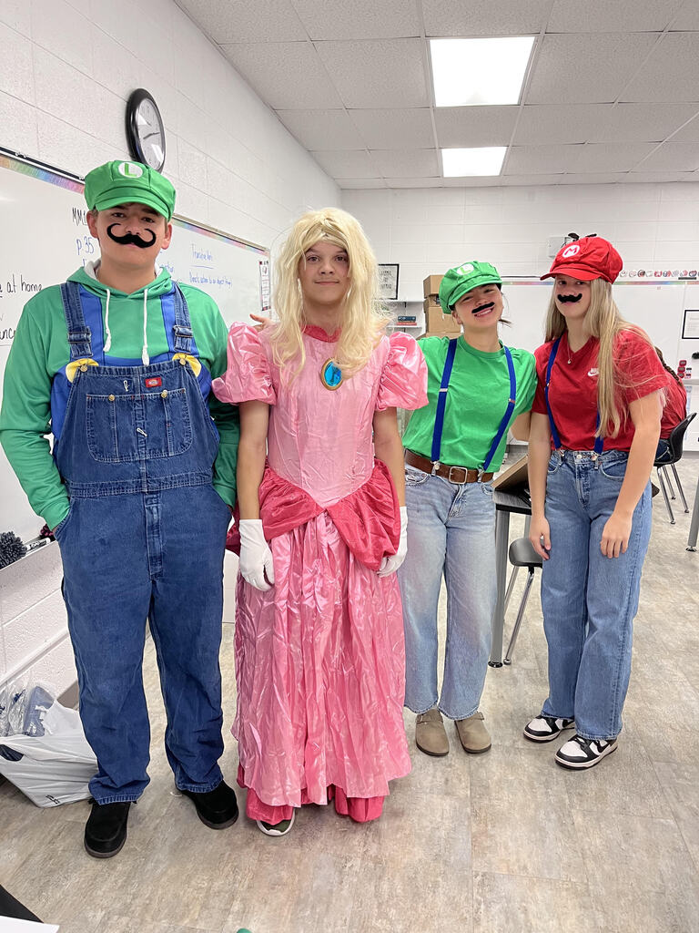 Four students pose in a classroom wearing video game-themed costumes. Two wear green outfits with blue overalls and green caps, while another wears a pink princess-style dress and blonde wig. The fourth wears a red shirt, blue jeans, a red cap, and a black mustache. A whiteboard, desks, and classroom supplies are visible in the background.