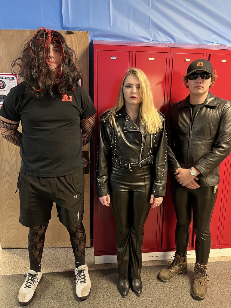 Three students pose in front of red school lockers wearing costumes. The student on the left wears a long black-and-red wig, black clothing, fishnet sleeves and tights, and white sneakers. The student in the center wears a black leather jacket with studs, black pants, and pointed boots. The student on the right wears sunglasses, a black cap, a brown leather jacket, black pants, and brown boots. 
