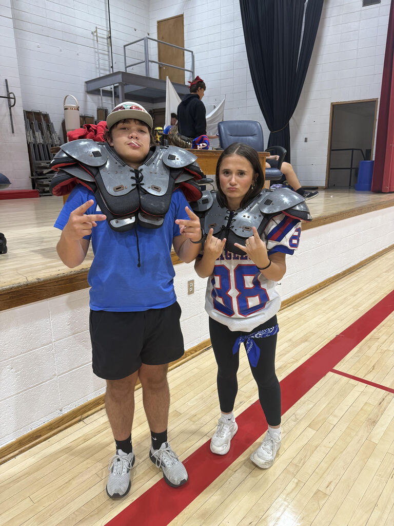 Two students pose on a school gym floor wearing football-themed costumes. Both wear football shoulder pads over Eagles jerseys, with the student on the left wearing a blue shirt, black shorts, and a cap, and the student on the right wearing a white Eagles jersey, black leggings, and a blue bandana. They make hand gestures toward the camera, with a stage and gym equipment visible in the background.