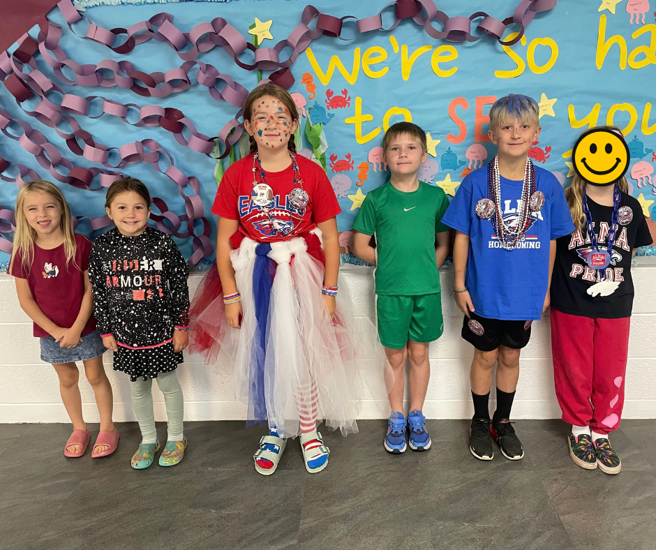 Five children pose together in front of a colorful school spirit backdrop decorated with paper chains, stars, sea creatures, and the words “We’re so happy to see you.” The children are wearing colorful outfits and school spirit accessories, including beads, a tutu, and face paint.