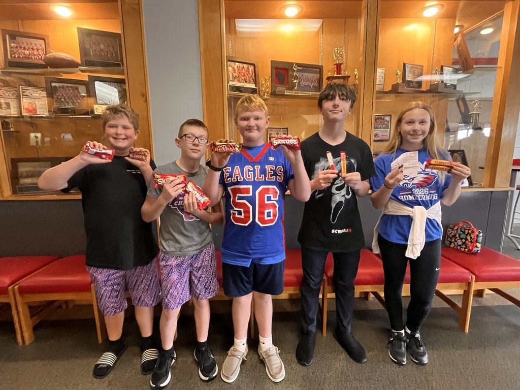 ive students pose together in front of a school trophy display while holding candy bars. They are wearing casual clothing and school spirit gear, including an Eagles jersey and a Homecoming shirt.