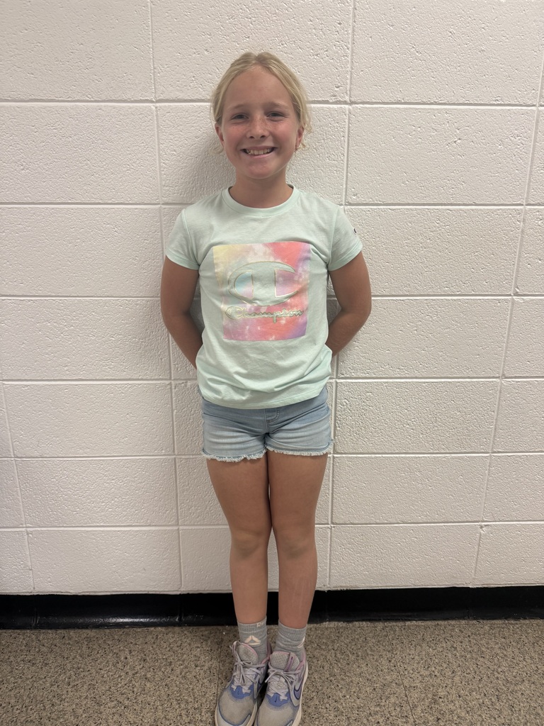A smiling young girl stands in front of a white brick wall, wearing a light green Champion T-shirt, denim shorts, gray socks, and sneakers.