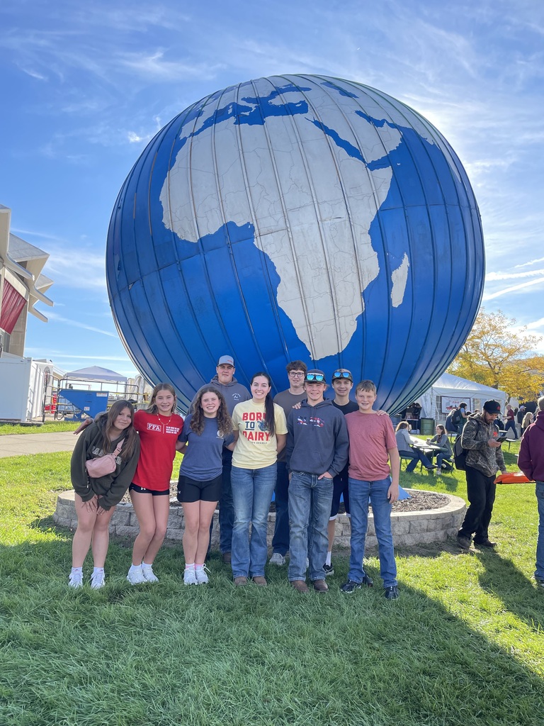 A group of nine teenagers posing together on a grassy lawn in front of a large blue globe sculpture showing the continent of Africa under a clear blue sky.