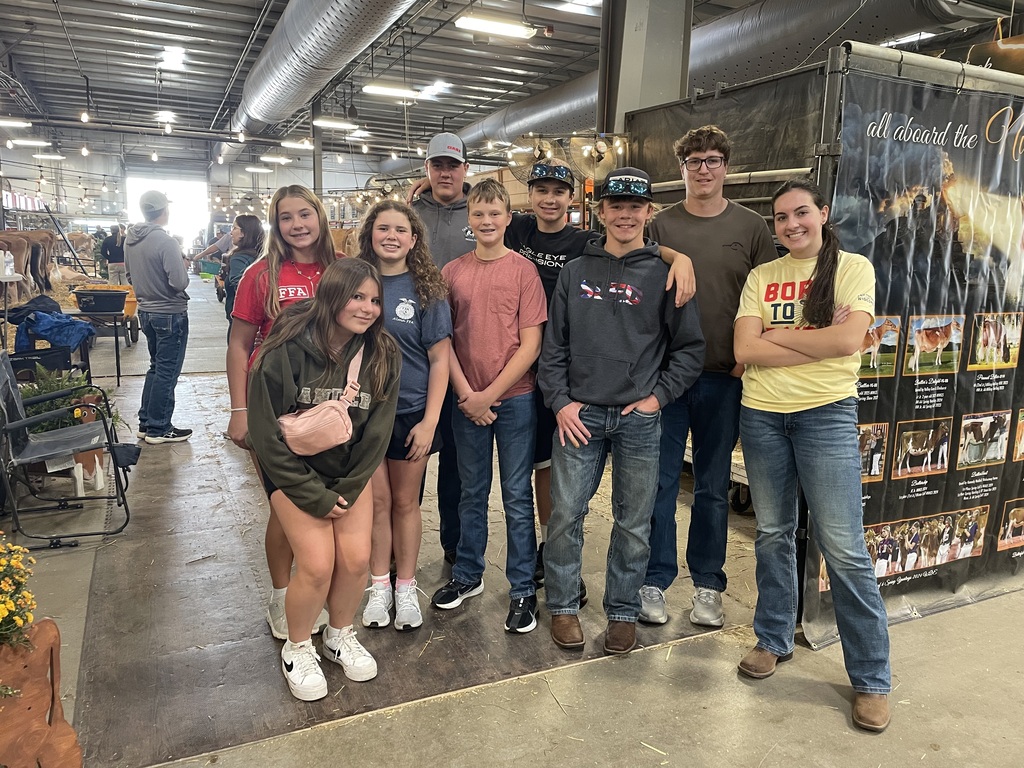 A group of eight young people pose together for a photo in a large livestock barn or agricultural pavilion. The group is casually dressed in t-shirts (including FFA and "Born to Dairy" logos), hoodies, jeans, and baseball caps. In the background, cattle stalls line the aisle to the left, and a large promotional banner displaying cattle photos stands on the right.