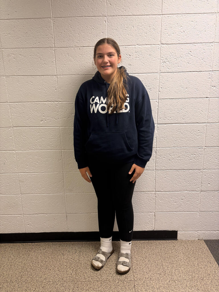 A young girl standing in front of a white wall wearing a dark blue hoodie, black tights, and socks & sandals.