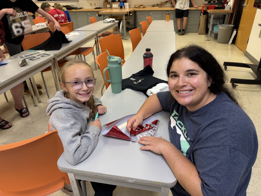 A young girl and an adult woman smile for the camera while working on a sports decal at a classroom table. Other students are working on shirts and decals in the background.