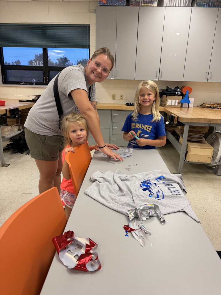 An adult and two young girls work together at a classroom table on sports shirt decals. The table has a gray sweatshirt with a blue design, vinyl scraps, and crafting materials.