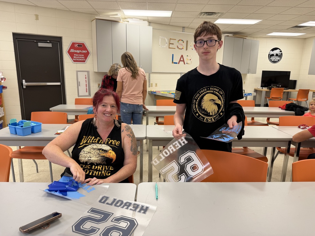 A student and an adult sit at a classroom table while working on blue and white sports decals. The student holds a large “Herold” number 23 decal, while other people work in the background.