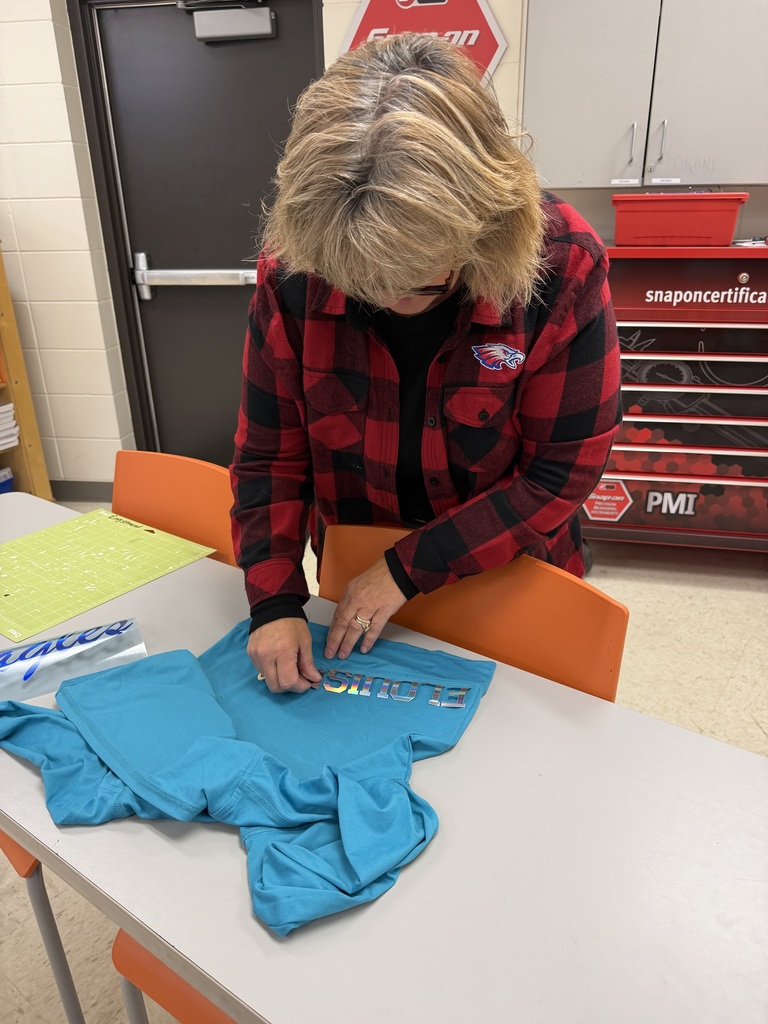 An adult wearing a red and black plaid shirt works on applying a colorful decal to a turquoise shirt at a classroom table. A cutting mat and other materials are nearby.