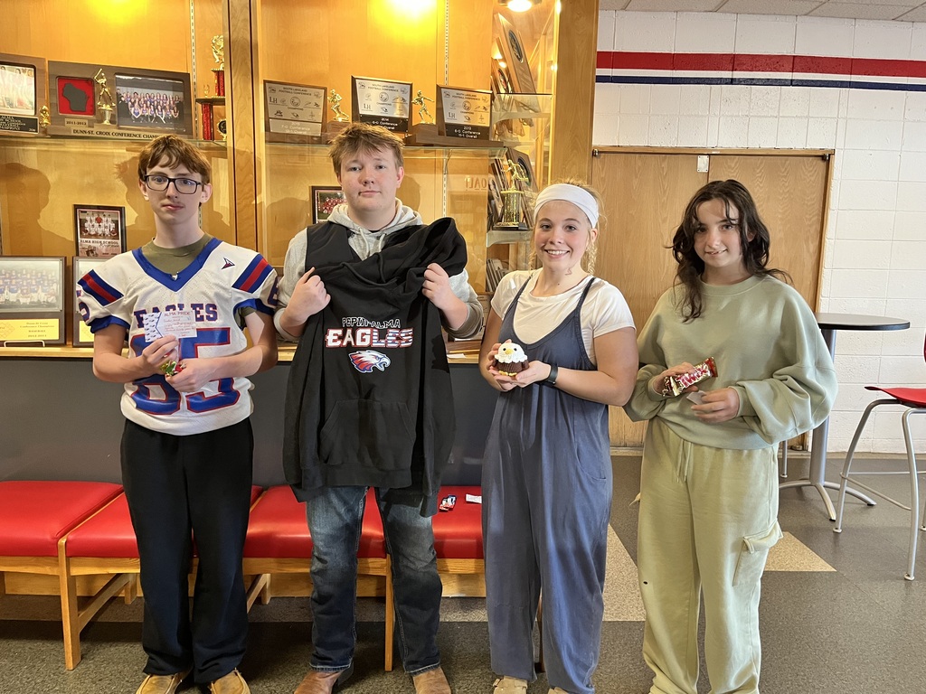Four students pose together in a school hallway, with two students holding an Eagles sweatshirt and sports jersey. The group stands in front of a display case filled with school trophies, plaques, and team photos.