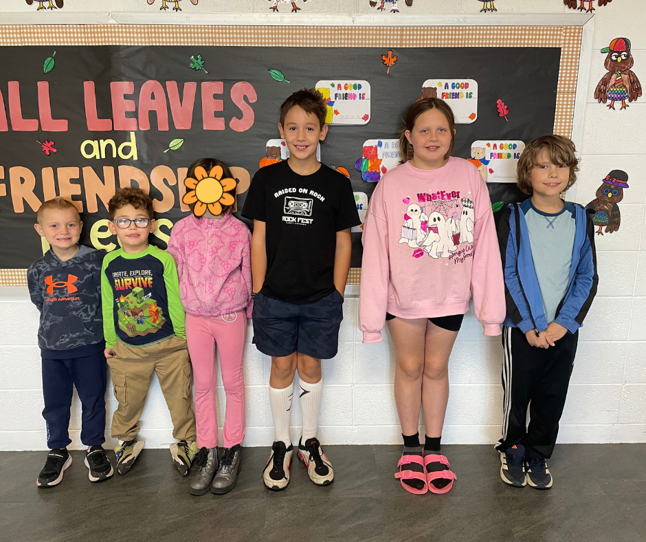 Six elementary school students stand together in front of a colorful “All Leaves and Friends” bulletin board. The students are smiling and wearing a variety of colorful outfits, with student artwork displayed on the wall behind them.