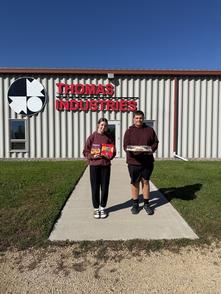 Students standing outside of a building labeled Thomas Industries. The students are holding a meat and cheese tray as well as crackers.