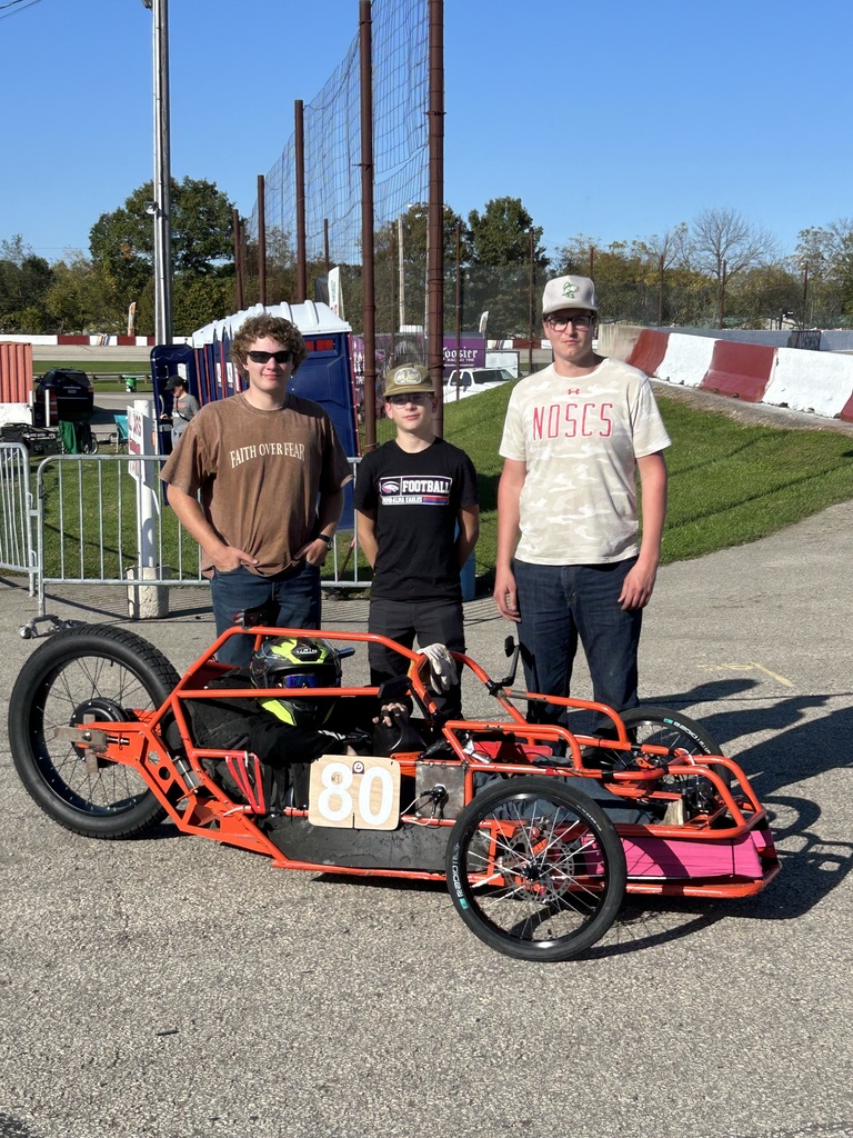 Three people pose beside a bright orange three-wheeled racing cart with the number 80 at an outdoor track. One person is seated inside the cart wearing a helmet, while the other two stand behind it. A tall safety fence, track barriers, and a clear blue sky are visible in the background.