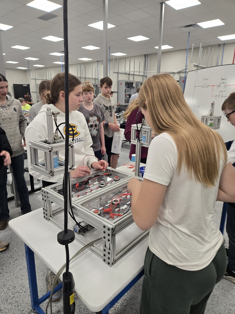 Students examine a tabletop engineering prototype with metal frames and wiring in a bright classroom, creating a focused, hands-on STEM learning atmosphere.