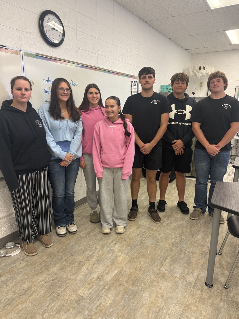 Seven students pose for a group photo in a classroom, standing in front of a whiteboard. They are dressed casually in hoodies, shirts, jeans, and athletic wear, with desks and classroom supplies visible around them.