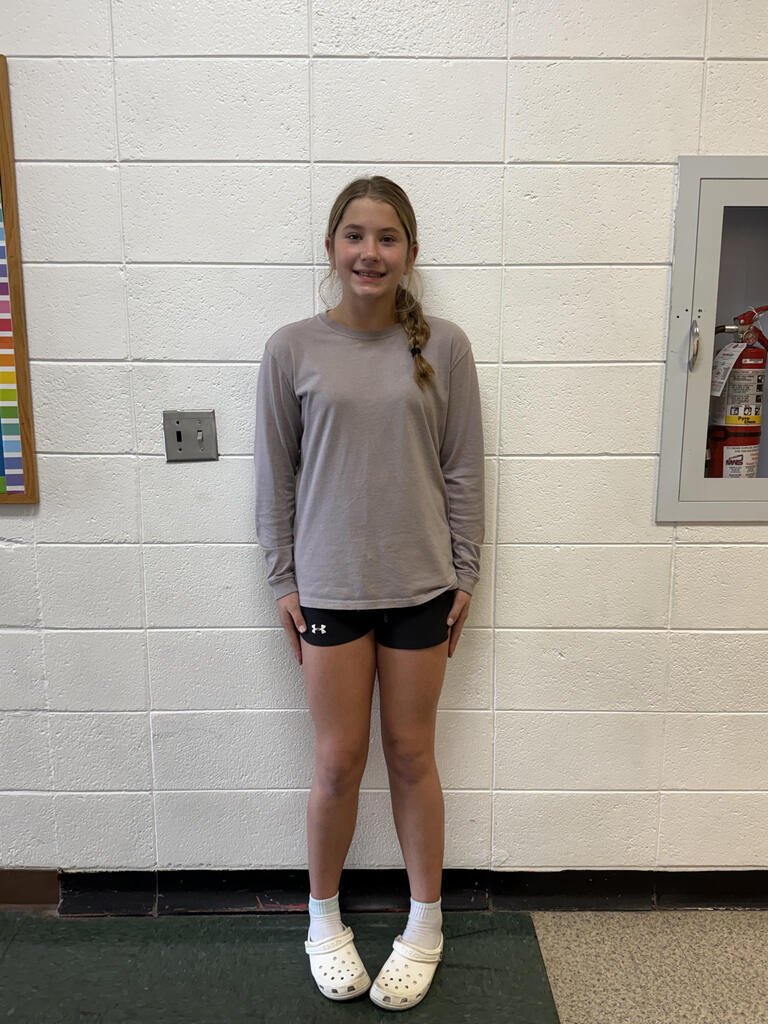 A student stands smiling in front of a white cinderblock wall in a school hallway, wearing a gray long-sleeve shirt, black athletic shorts, white socks, and white Crocs.