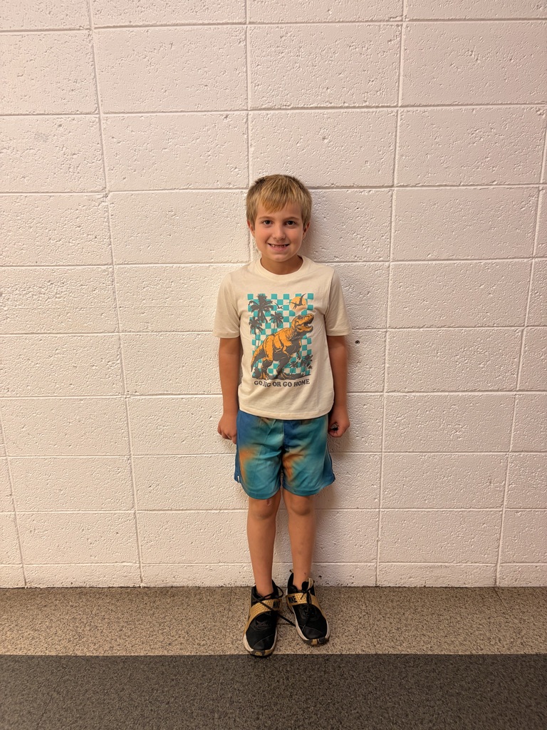 A smiling young boy stands against a pale tiled wall, wearing a colorful graphic T-shirt, turquoise shorts, and black sneakers; the portrait feels cheerful and casual.