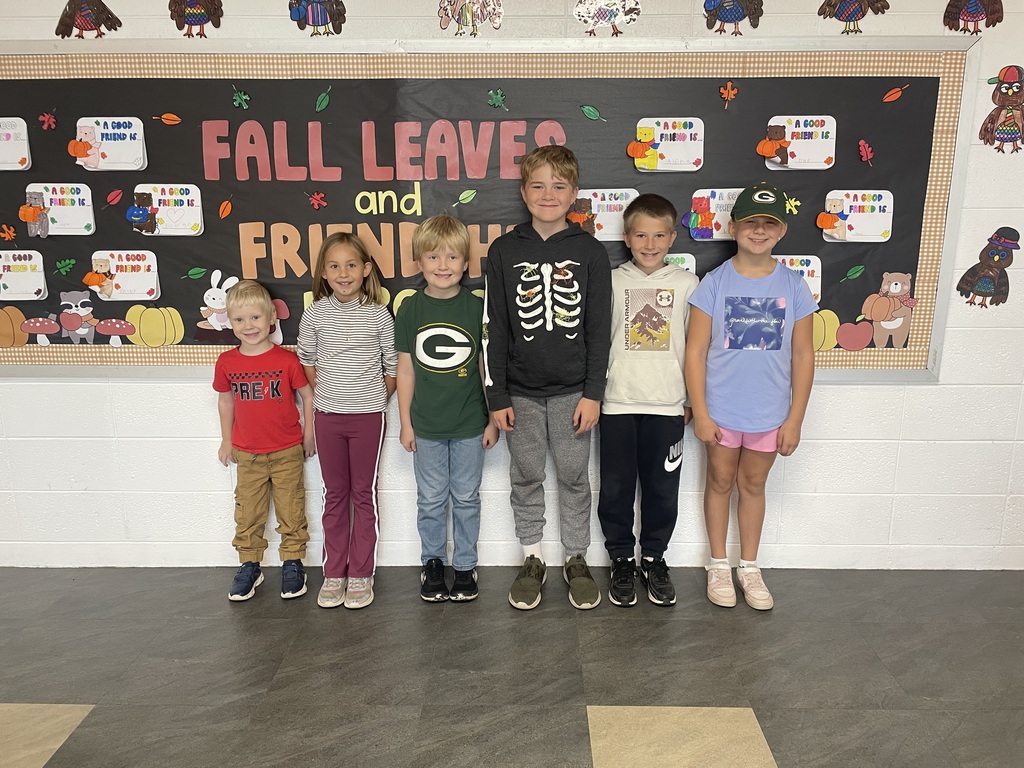 Six elementary-age children smile in a school hallway before a colorful “Fall Leaves and Friends” bulletin board, wearing casual clothes and a skeleton-print shirt.