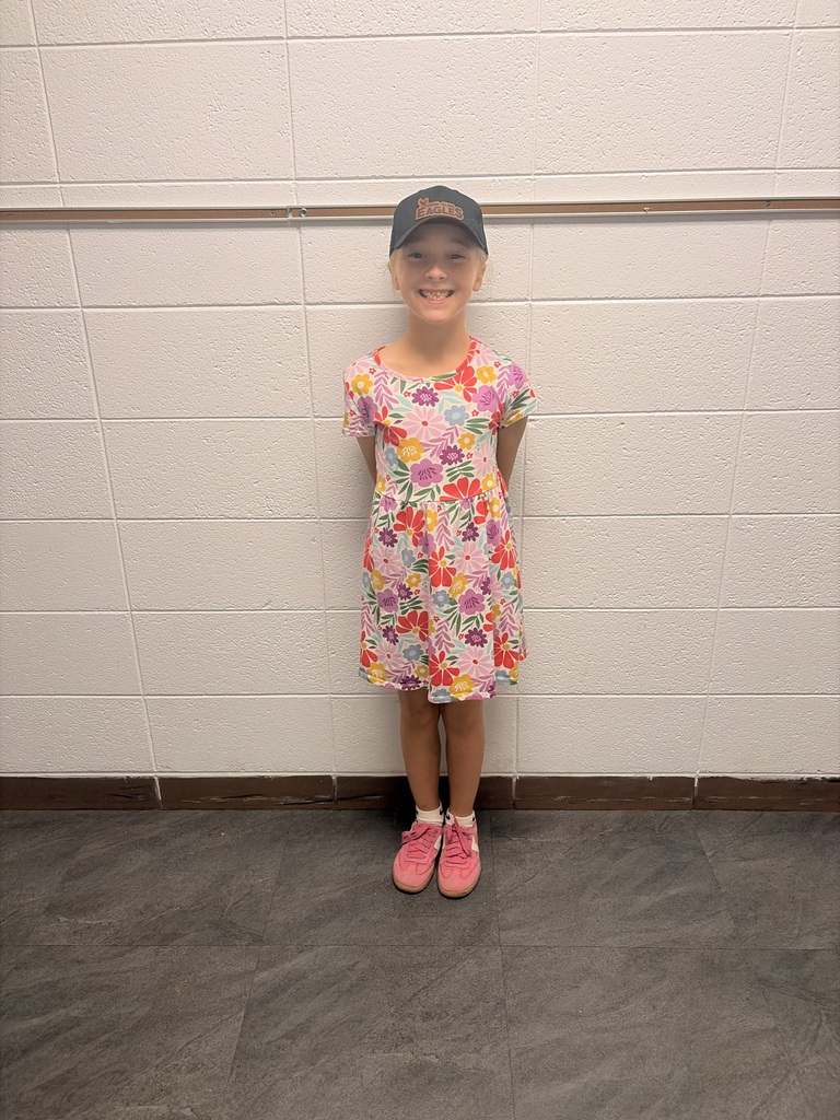 A smiling child stands against a tiled hallway wall in a bright floral dress, black cap, and pink sneakers, creating a cheerful, confident mood.