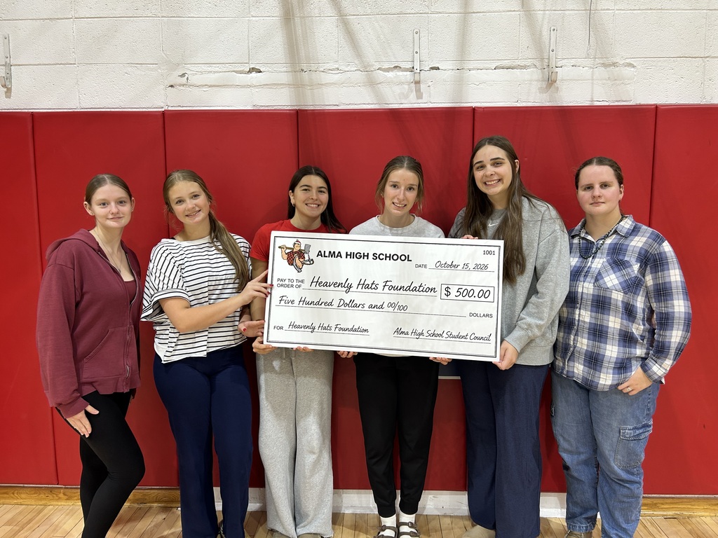 Six students pose against red gym mats, holding a $500 check from Alma High School to Heavenly Hats Foundation—a cheerful group donation presentation.