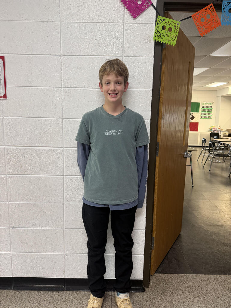 A student stands smiling in a school hallway beside an open classroom door. He wears a green T-shirt over a blue long-sleeve shirt, black pants, and tan shoes. Colorful decorative skull banners hang above the doorway, and desks and chairs are visible inside the classroom.