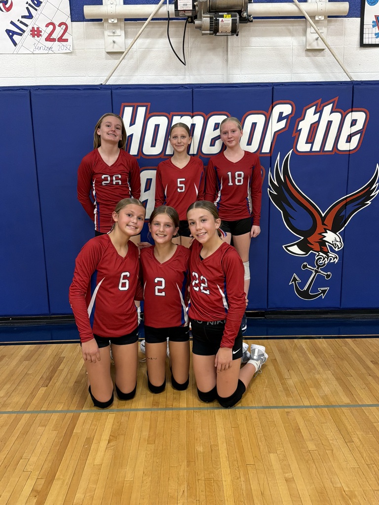 Six young volleyball players pose together in red jerseys and black shorts on a gym floor. Three players stand in the back row and three kneel in the front, smiling in front of blue wall padding featuring the school's eagle logo.