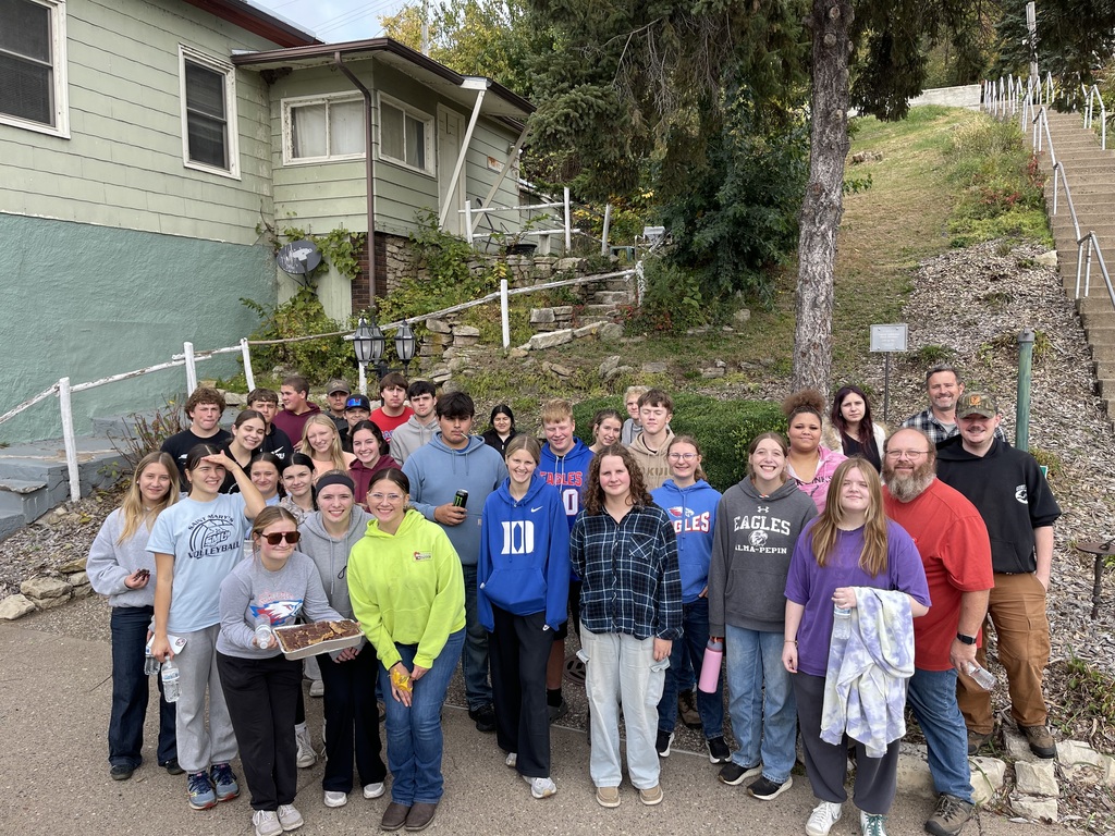 A large group of students and adults pose together for a photo outdoors on a sidewalk at the base of a hillside. Behind them are a green building, stone retaining walls, trees, and a long concrete staircase with white railings. Several people are smiling, and some are holding water bottles, snacks, and personal belongings.