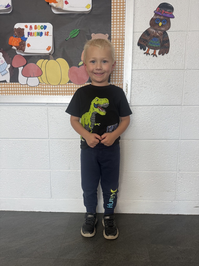 A smiling young child with blonde hair stands in a classroom, wearing a black dinosaur graphic T-shirt, navy blue joggers, and black sneakers. Behind him is a colorful fall-themed bulletin board with pumpkins, mushrooms, and student artwork, along with a handmade owl decoration on the white brick wall.