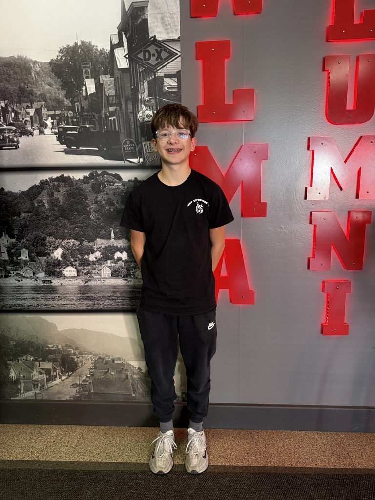 A smiling teenager with glasses and braces stands with his hands behind his back, wearing a black T-shirt, black Nike joggers, gray socks, and white sneakers. He poses in front of a wall featuring large red illuminated letters and black-and-white historical photographs of a town.
