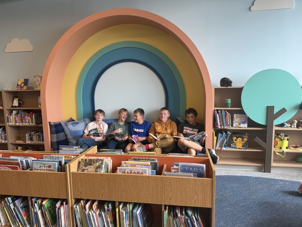Five boys sitting together under a rainbow mural all with books in their hands.